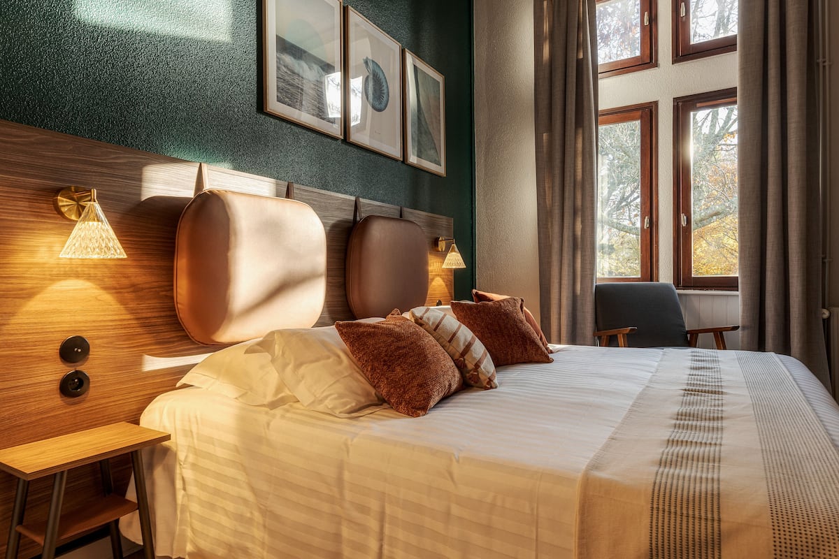 A neatly made bed with a headboard, bedside lamps, and a view of autumn trees through large windows.