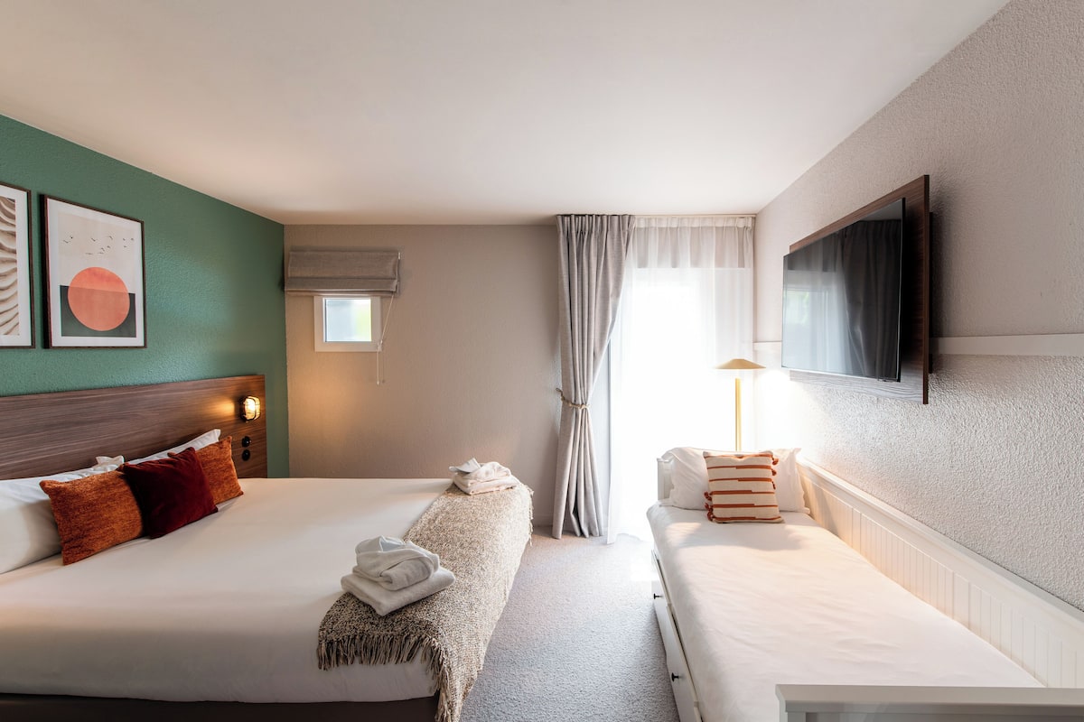A hotel room with two beds, a TV mounted on the wall, a window with curtains, and artwork on the wall.