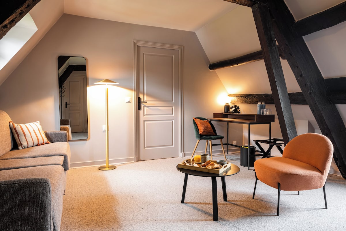 A cozy loft bedroom with a sloped ceiling, a large mirror, a modern sofa, an orange armchair, and a breakfast tray on a small table.