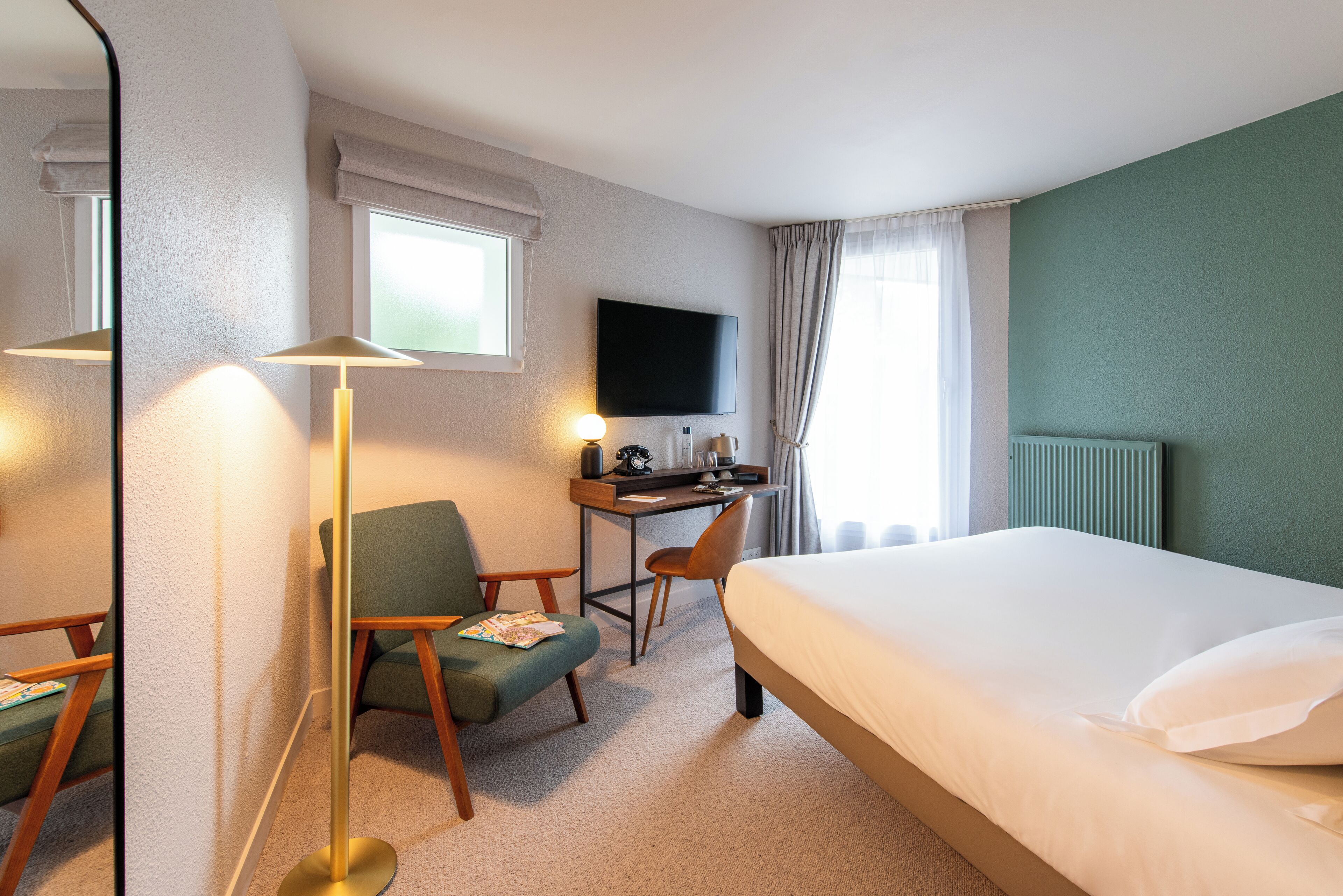 A hotel room with a bed, a desk, a chair, a TV, and a floor lamp.