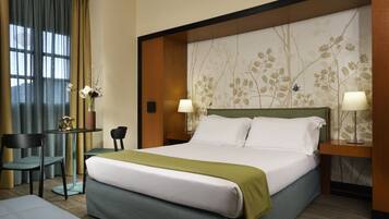 A hotel room with a large bed, two chairs, a small table, and a decorative wall panel.