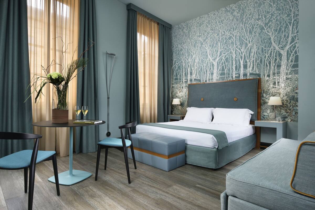 Una habitación de hotel moderna con una cama grande, una zona de estar con mesa y sillas, una mesita auxiliar y una planta decorativa.
