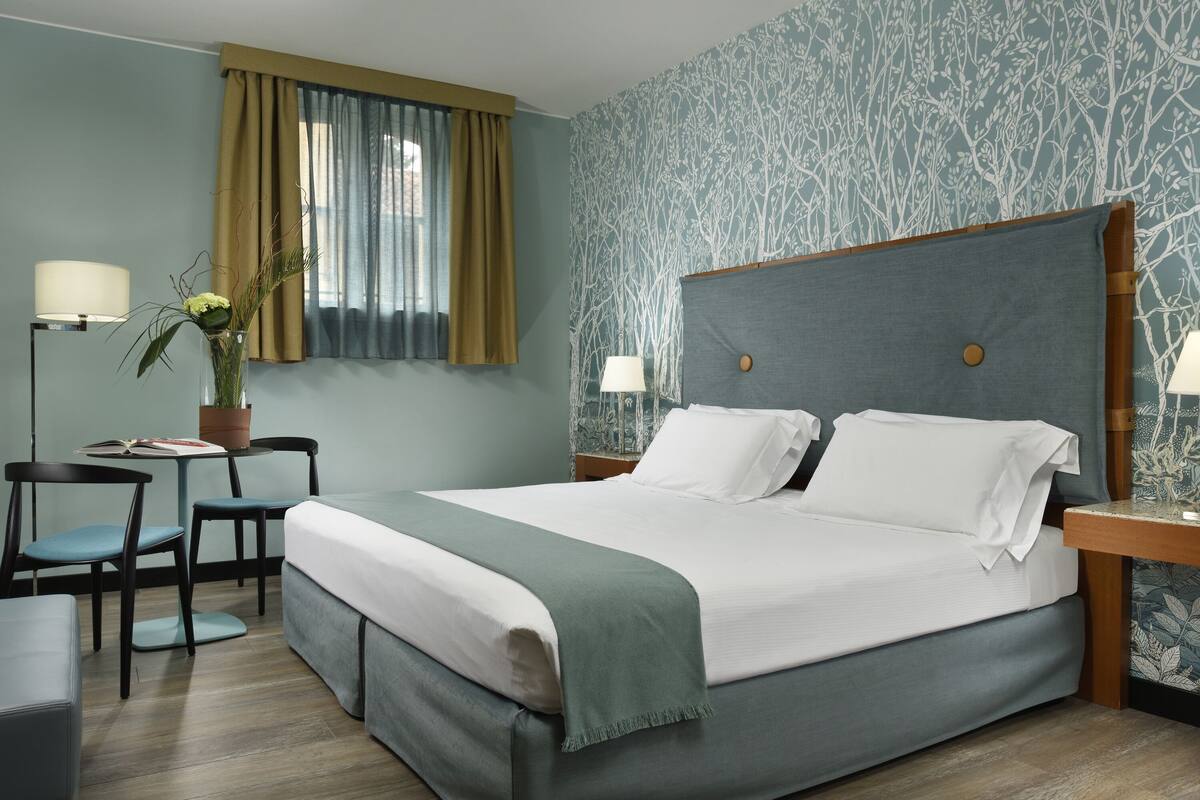 Habitación de hotel ordenada con una cama grande, una mesita con una lámpara, una silla y una planta en maceta.