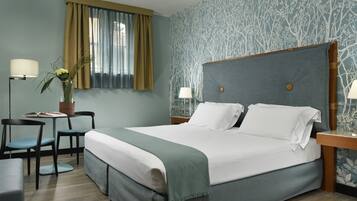 Habitación de hotel ordenada con una cama grande, una mesita con una lámpara, una silla y una planta en maceta.