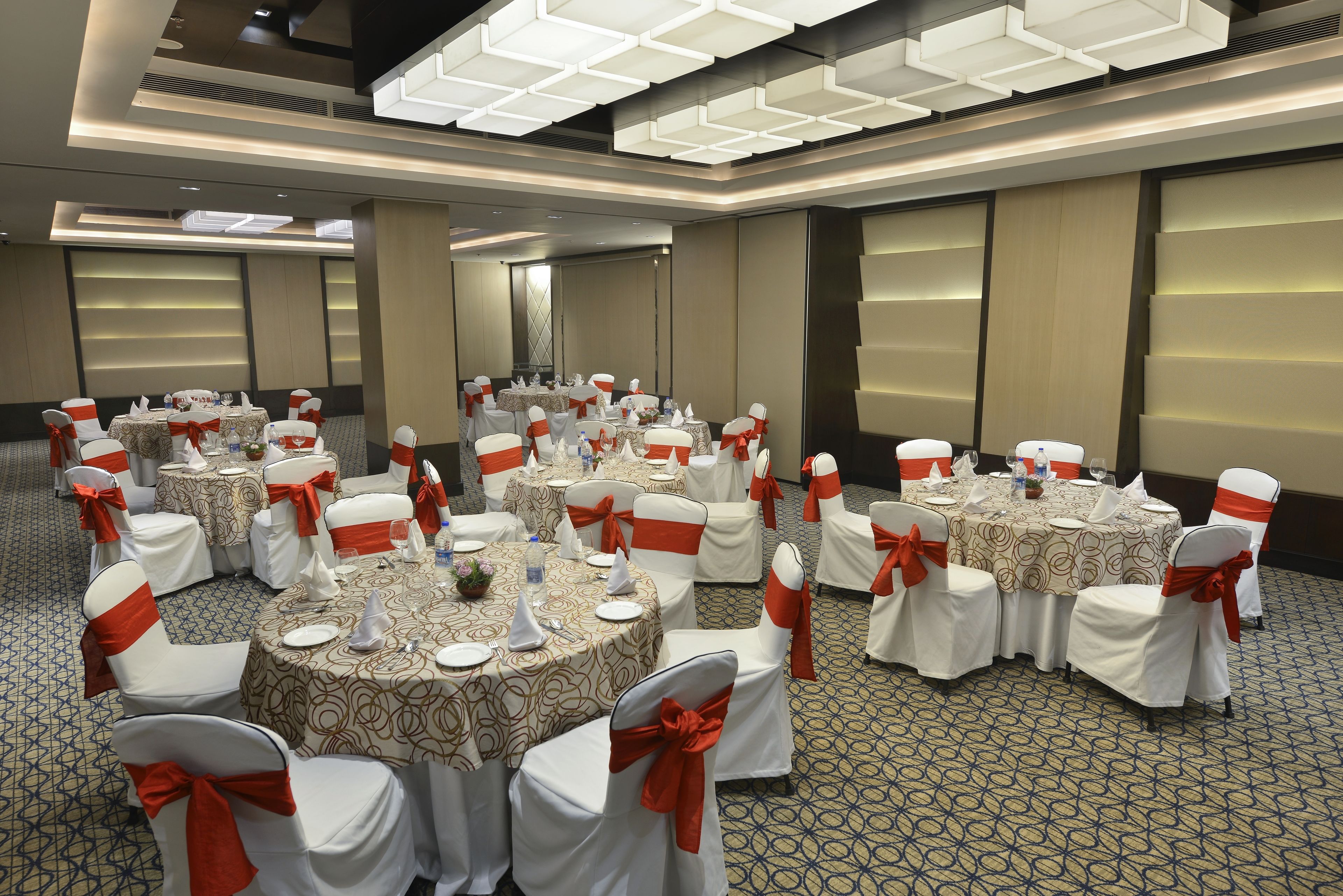 Un salón de banquetes con mesas redondas, sillas blancas con cintas anaranjadas y una alfombra estampada.