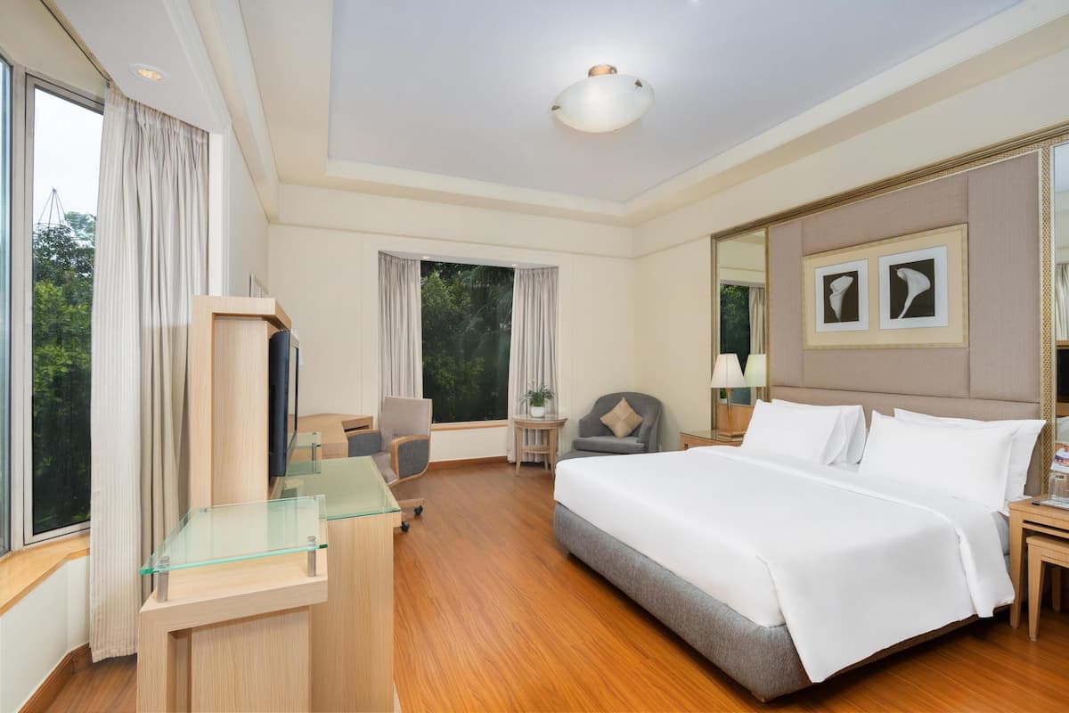 Ein Hotelzimmer mit einem großen Bett, einem Schreibtisch mit Glasplatte, einem Stuhl, einem kleinen Tisch mit Lampe und einem großen Fenster mit Vorhängen.