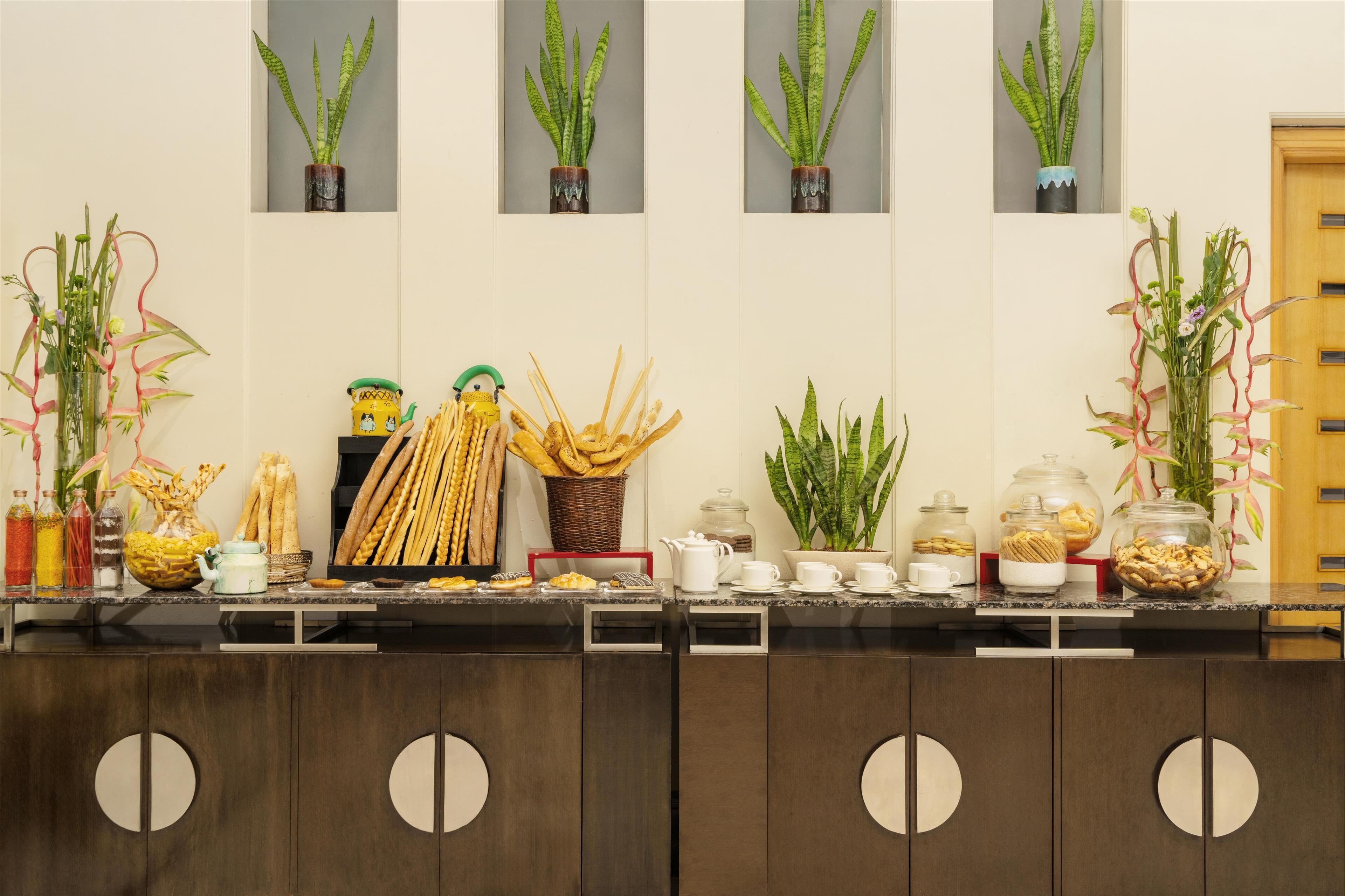 A buffet counter with various food items, including bread, pastries, and condiments. There are also several potted plants and decorative items on the wall.