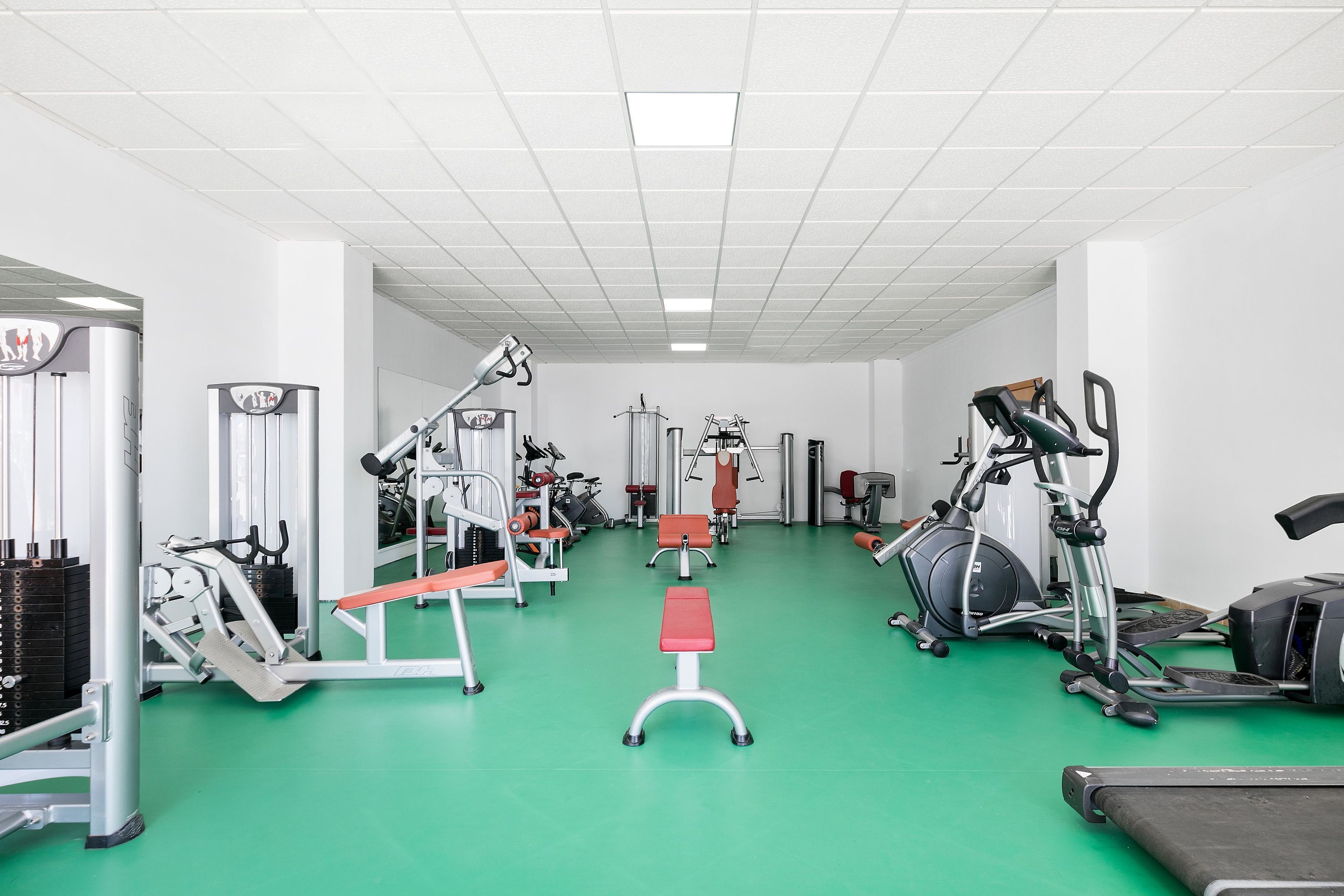 Ein Fitnessraum mit unterschiedlichen Trainingsgeräten, einem grünen Boden und weißen Wänden.