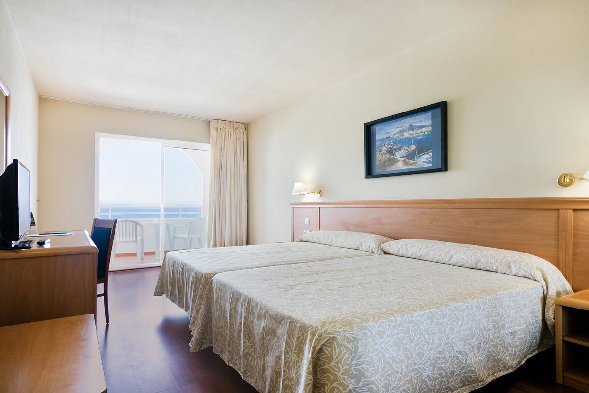 A hotel room with two beds, a desk, a chair, a TV, a balcony with a view of the sea, and a framed picture on the wall.