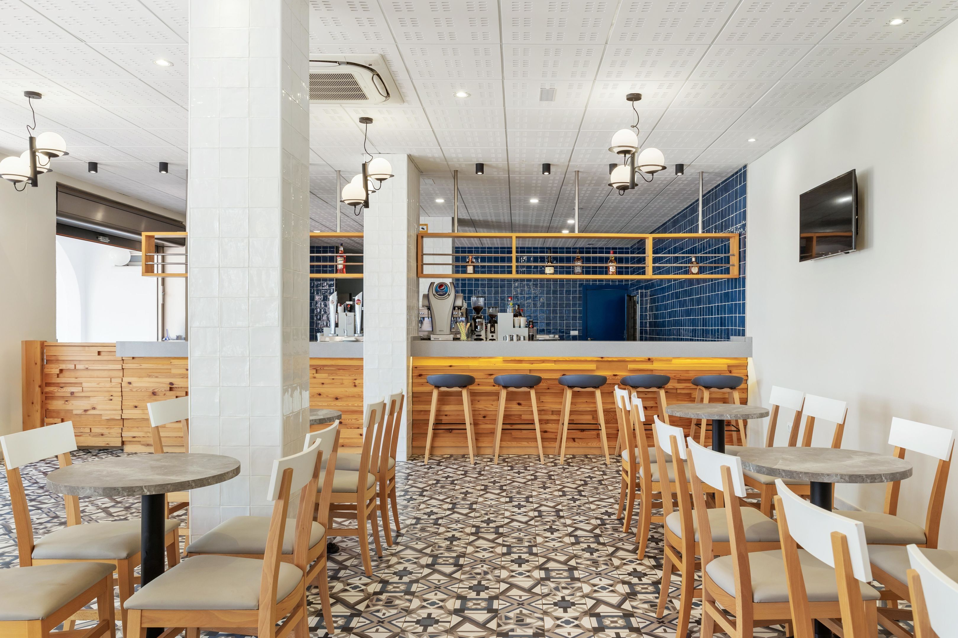 A modern restaurant interior with wooden tables and chairs, a patterned floor, and a blue-tiled counter area.