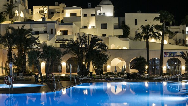 Vista nocturna d'una piscina amb reflexos de palmeres i edificis.