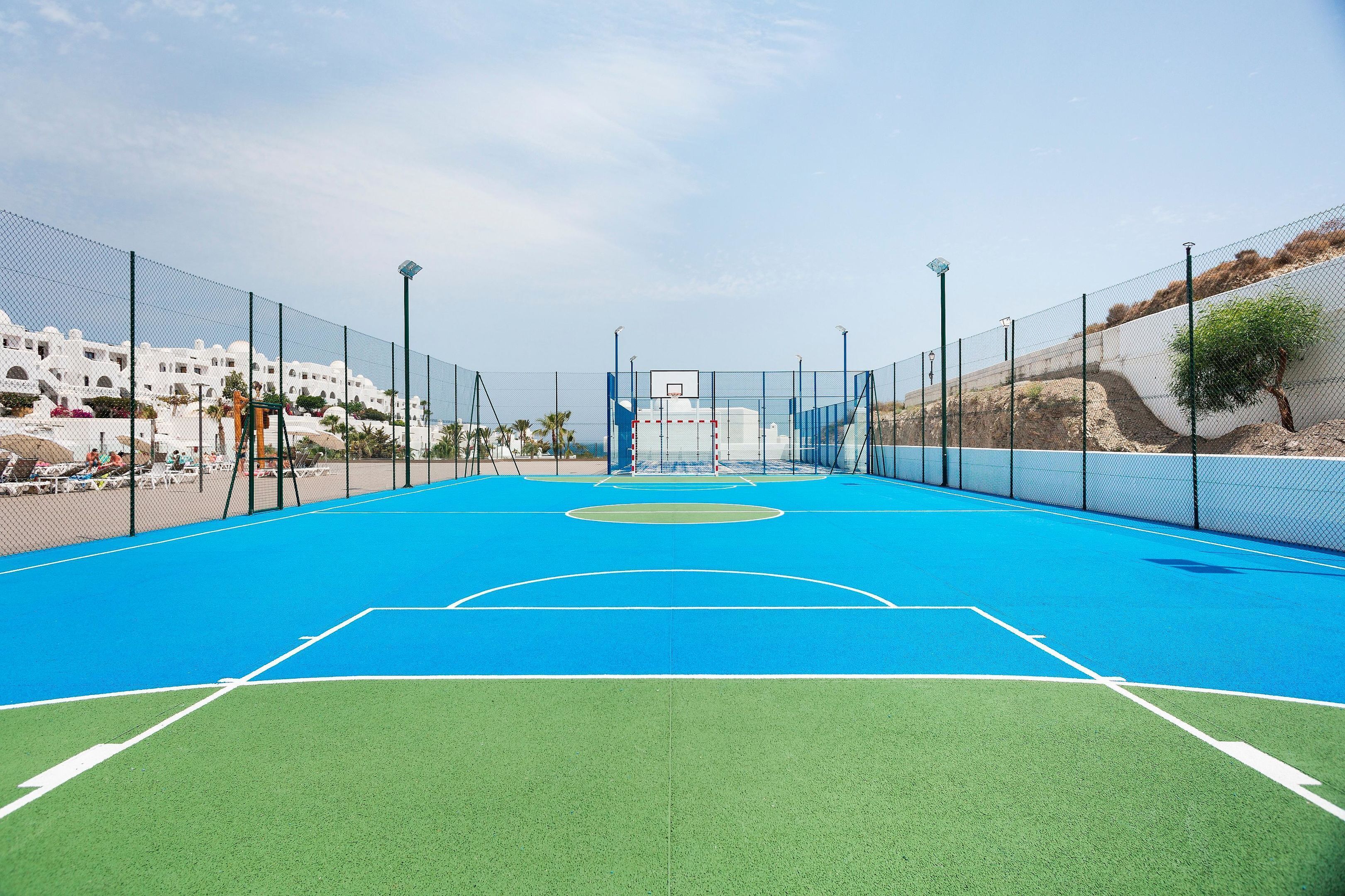 An outdoor basketball court with a blue playing surface, surrounded by a chain-link fence, and a white building in the background.