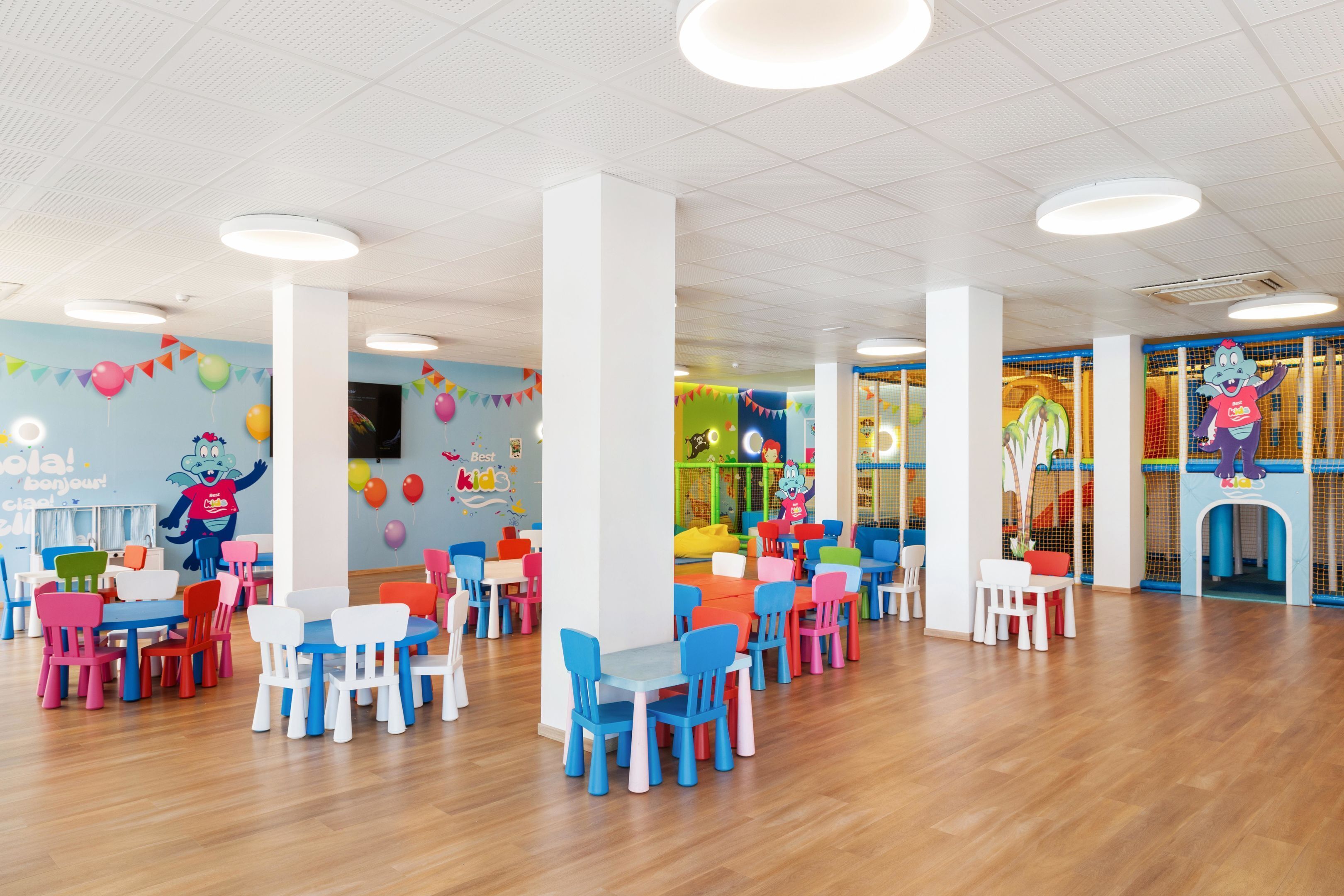 A brightly lit indoor space with colorful chairs and tables arranged in rows, a play area with a netted enclosure, and a mascot figure on the wall.