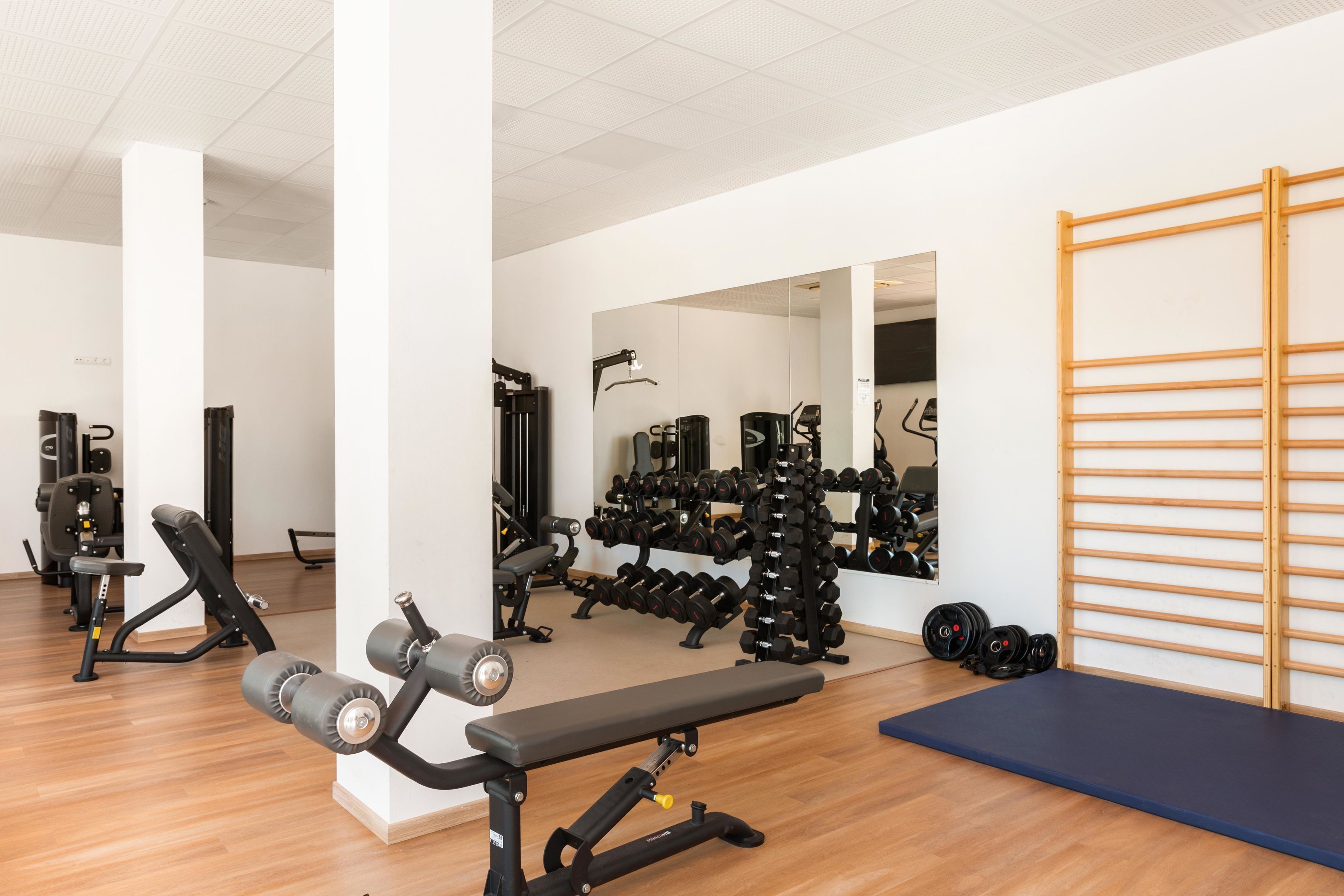 Ein Fitnessraum mit unterschiedlichen Trainingsgeräten, einer Bank und einer Spiegelwand.