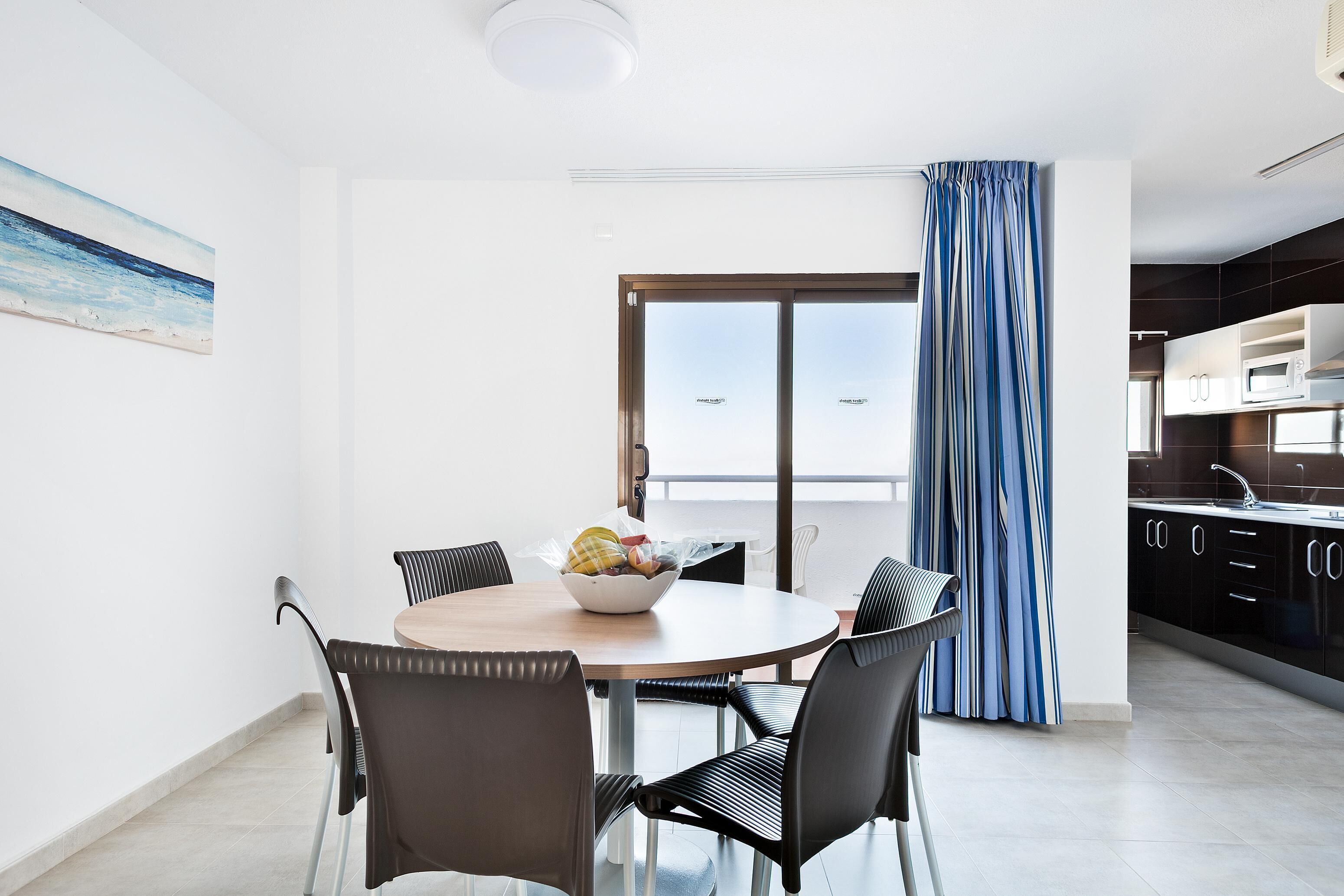 A modern kitchen with dark cabinets and a white countertop, a dining area with a round table and chairs, and a large window with a view of the sea.