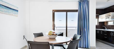 A modern kitchen with dark cabinets and a white countertop, a dining area with a round table and chairs, and a large window with a view of the sea.