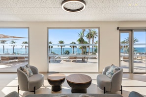 Une chambre d’hôtel moderne dotée d’une grande fenêtre donnant sur la plage et l’océan. On y trouve deux fauteuils et une table basse en bois.