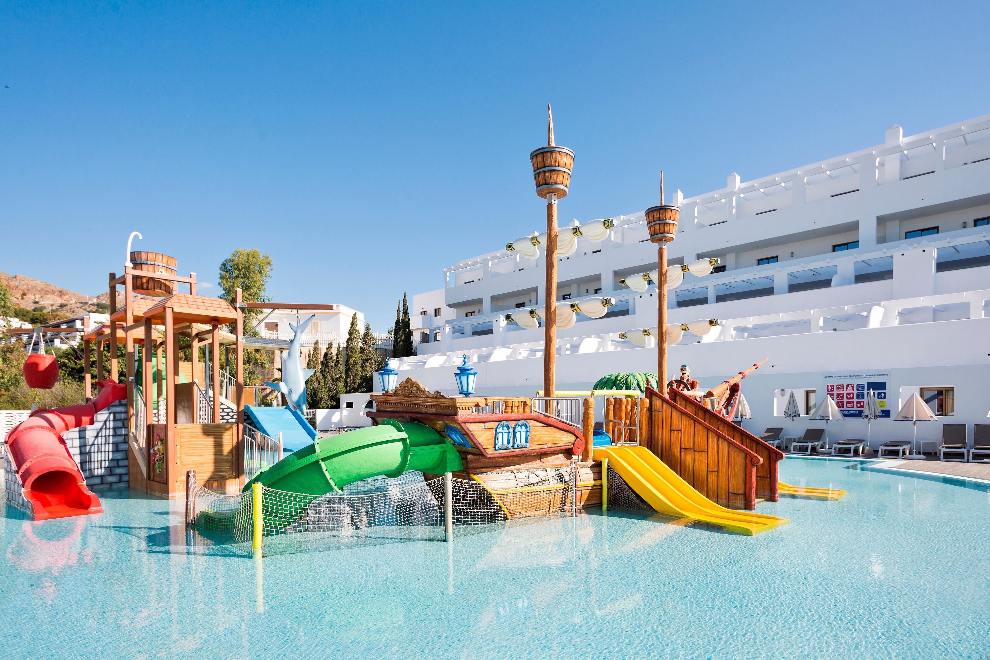 A water park with slides and play structures, a pool, and a white building with balconies in the background.