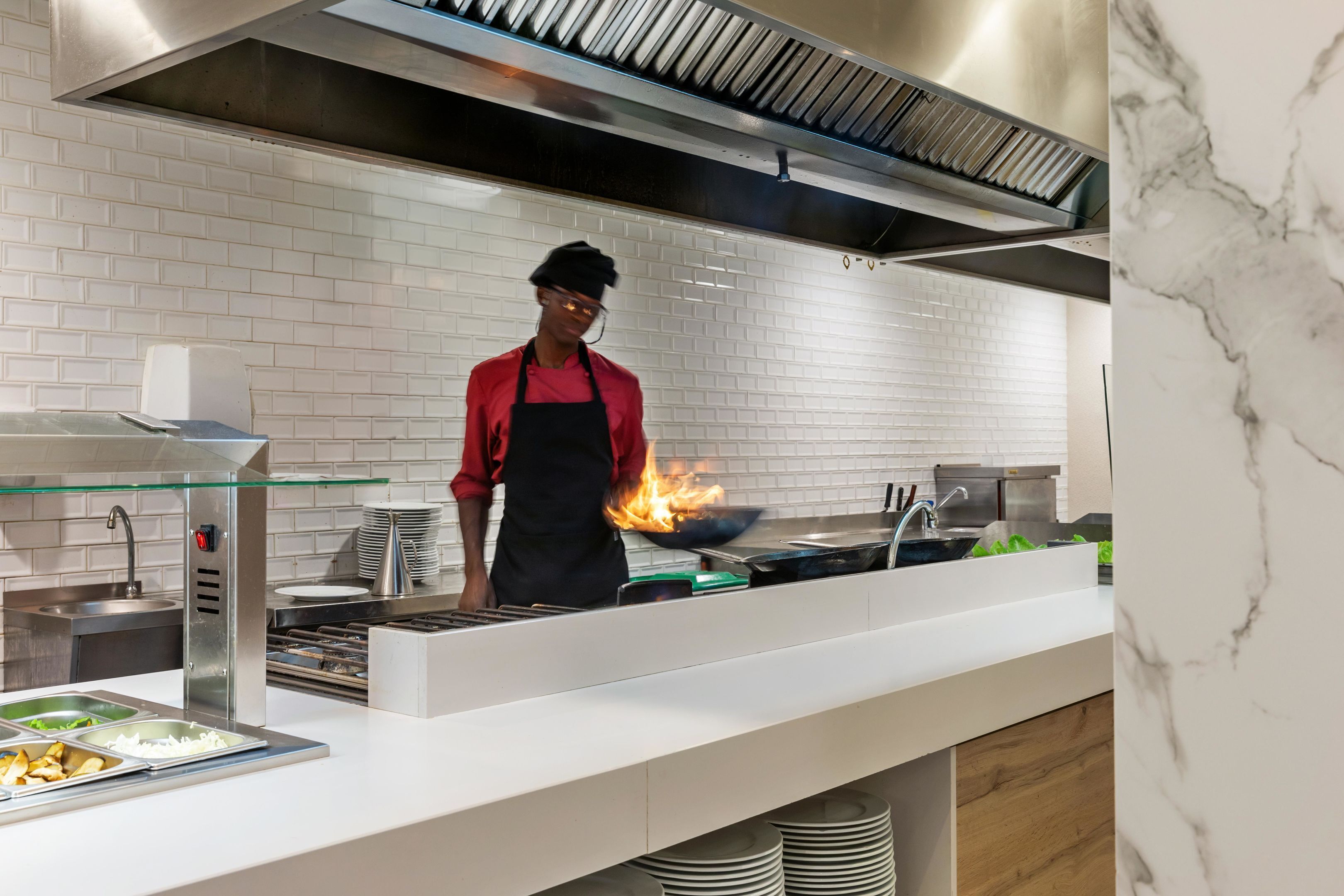 Una cocina moderna con una campana extractora de acero inoxidable, una olla china en la hornilla con llamas, y una zona de buffet con varios recipientes de comida.