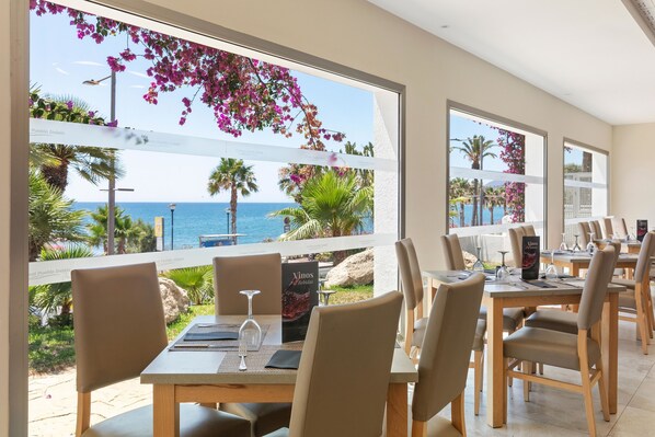A restaurant with tables and chairs, large windows with a view of the sea and palm trees, and a menu stand.