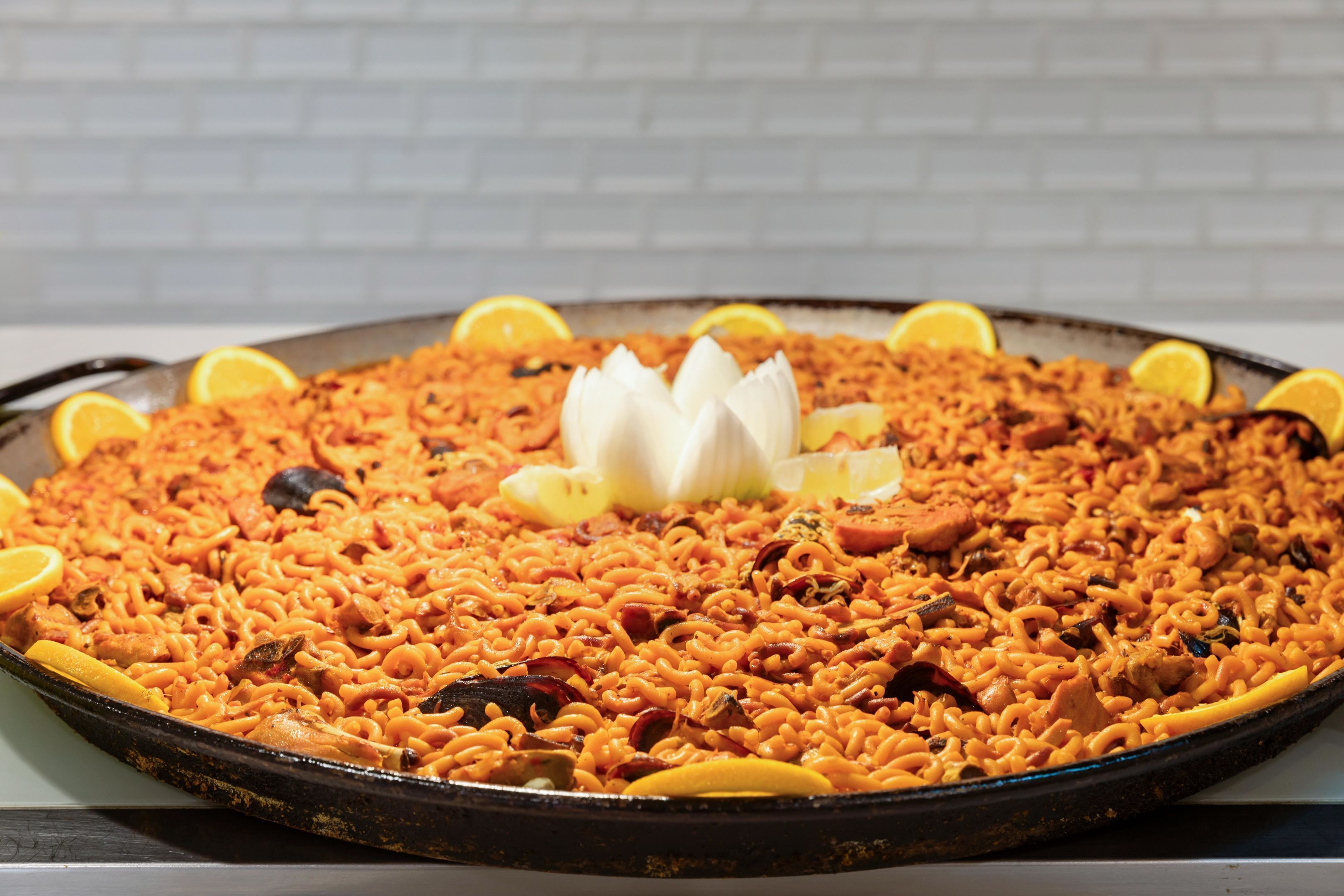 A large, round, black pan filled with a rice-based dish, garnished with lemon slices and a white flower.