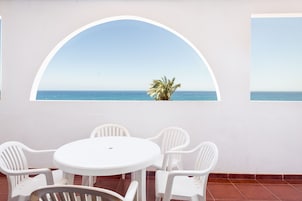 Una mesa y sillas blancas frente a una pared con un gran ventanal arqueado con vista al mar.