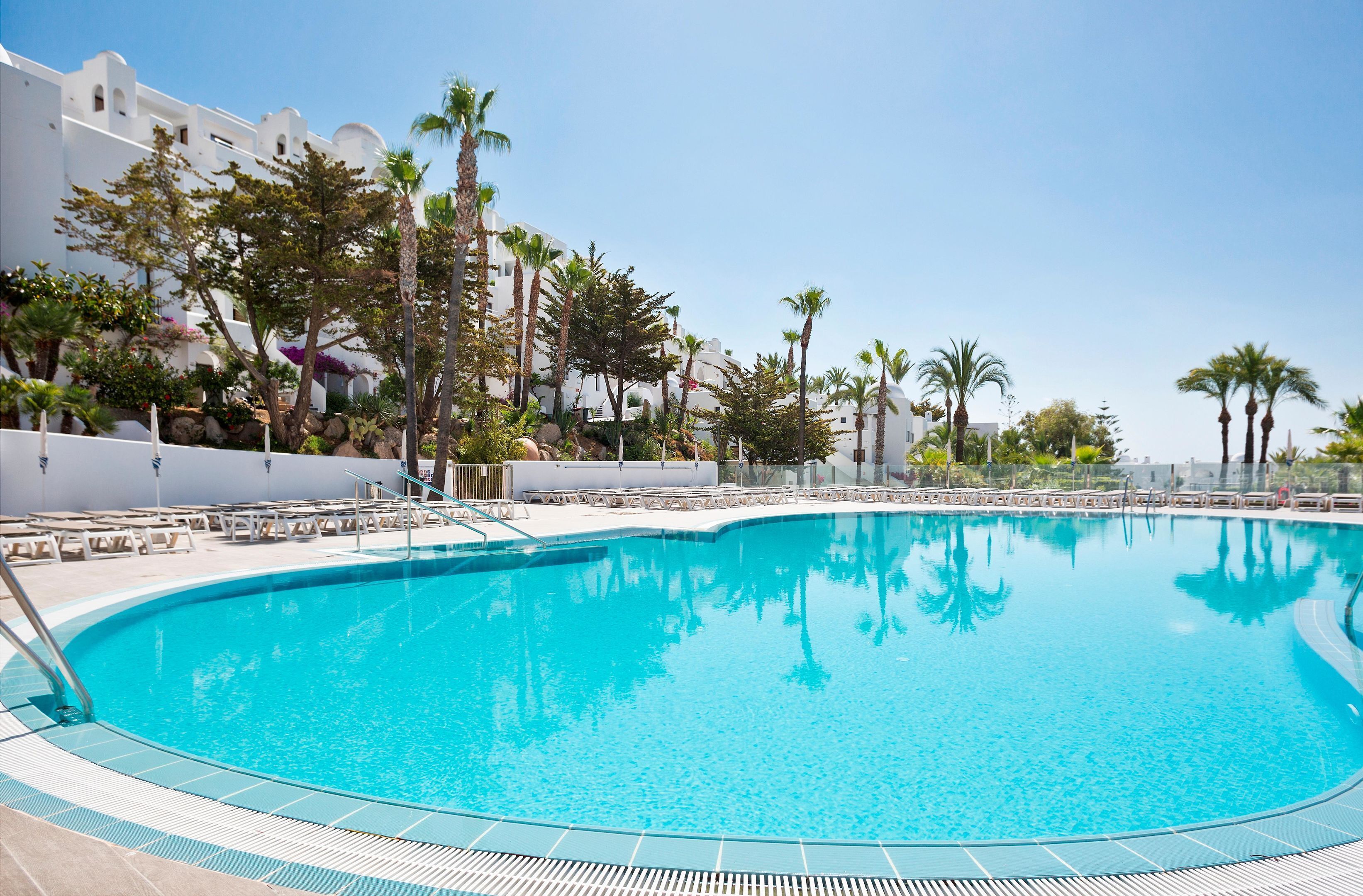 Una piscina azul cristalina rodeada de palmeras y edificios blancos con balcones.