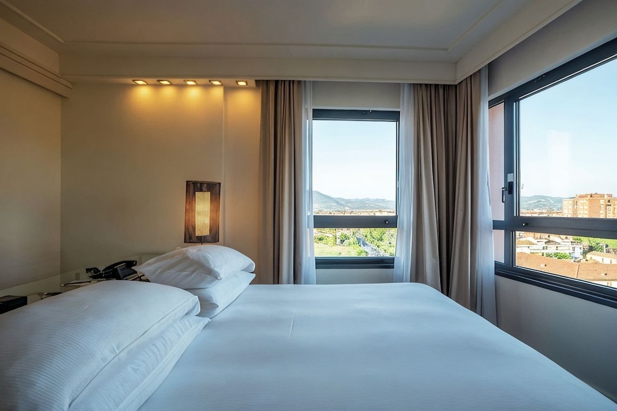 A hotel room with a large bed, a bedside table, a telephone, and a window with a view of the city.