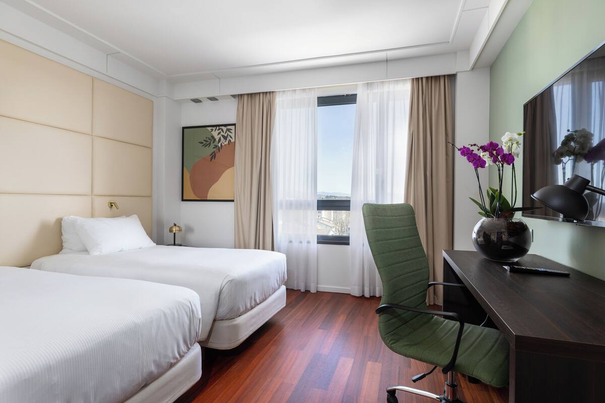 A hotel room with two beds, a desk with a chair, a desk lamp, and a window with curtains.