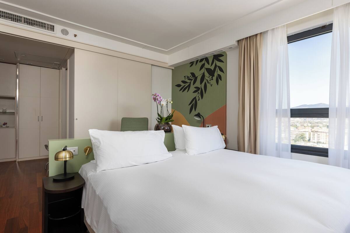 A hotel room with a large bed, a nightstand with a lamp, a green chair, and a view of the city through the window.