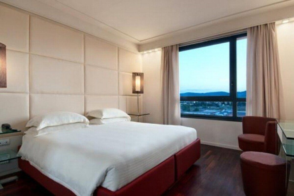 Habitación de hotel con cama, escritorio, silla y un ventanal con vistas.