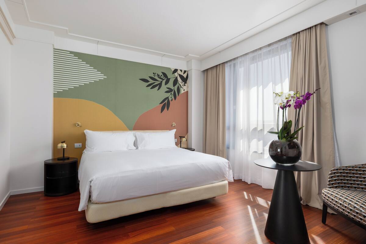 A modern hotel room with a large bed, a round side table, a small round table with a vase of flowers, and a patterned armchair.
