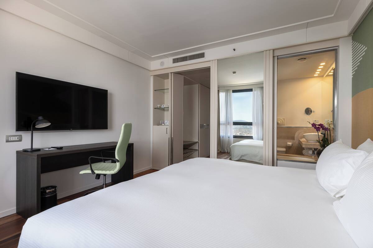 A hotel room with a large bed, a desk with a chair, a TV mounted on the wall, and a bathroom visible through an open door.