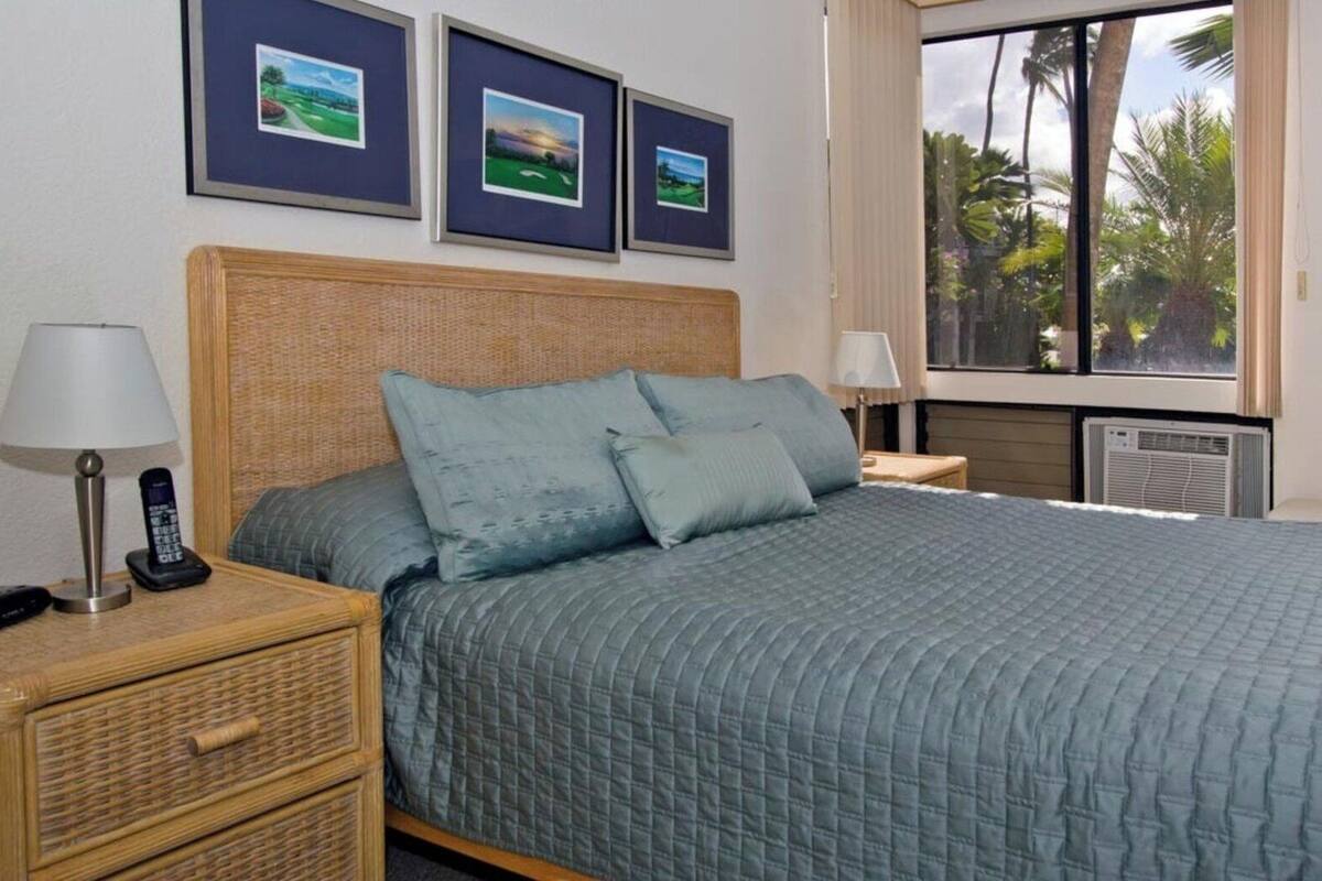 A neatly made bed with a blue quilt, a wicker nightstand with a lamp and phone, and framed pictures on the wall.
