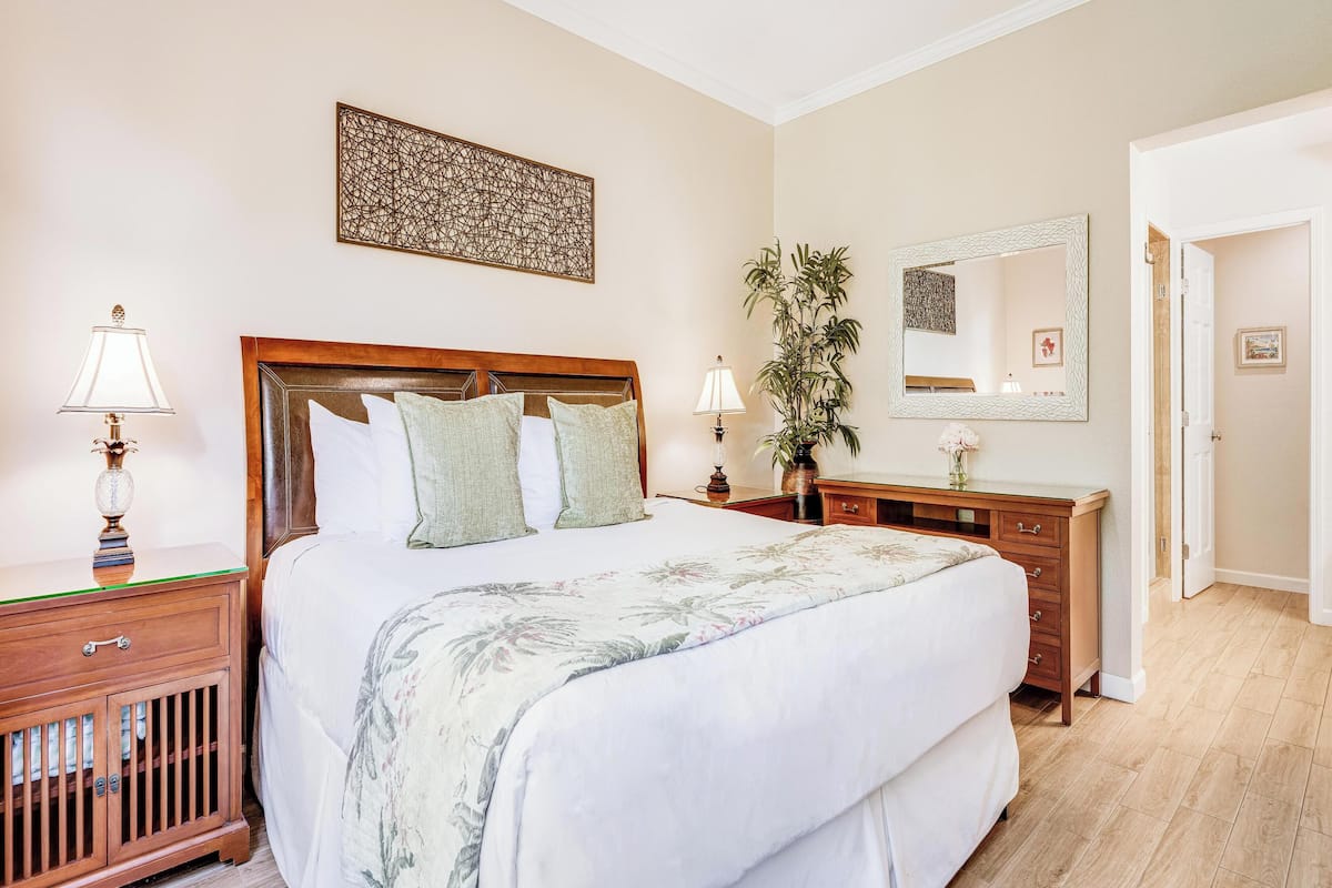 A bedroom with a wooden headboard, a large bed with floral bedding, a nightstand with a lamp, a dresser, a mirror, and a potted plant.