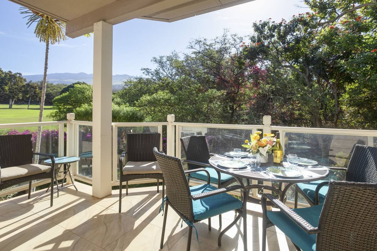 A covered outdoor seating area with wicker chairs and a glass table, overlooking a lush landscape with trees and a distant mountain range.