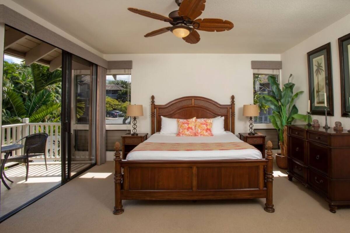 A bedroom with a wooden bed, a ceiling fan, a nightstand with a lamp, a dresser, and a sliding glass door leading to a balcony.