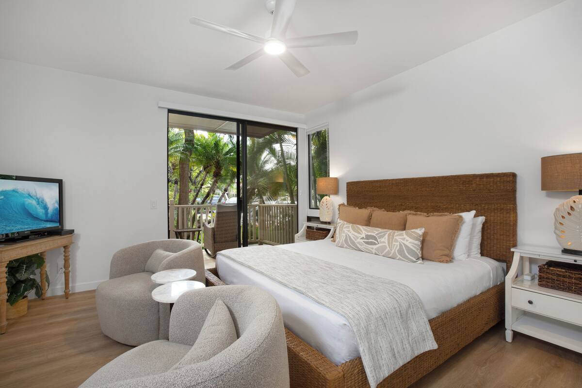 A bedroom with a large bed, a ceiling fan, a television, and a view of palm trees.