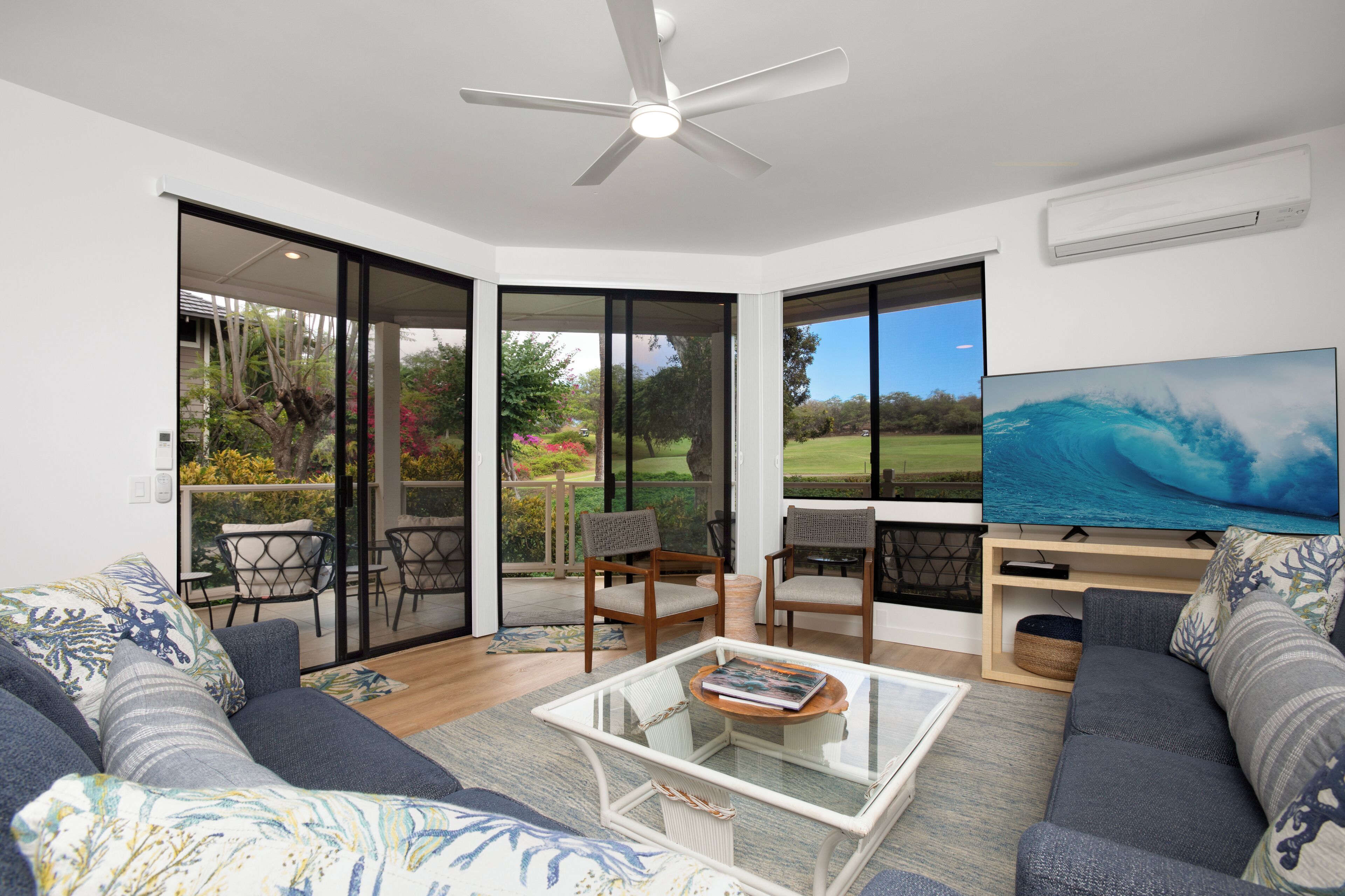 A living room with a large TV, a ceiling fan, and a view of a garden through glass doors.