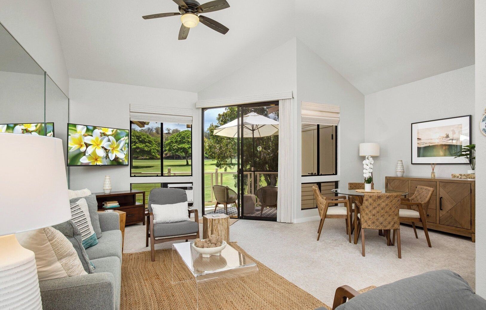 A spacious living room with a sofa, coffee table, and dining area. There is a large window with a view of a garden.