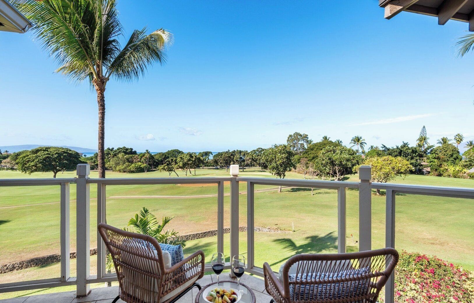 A balcony with wicker chairs, a table with a fruit bowl and wine glasses, and a view of a golf course.
