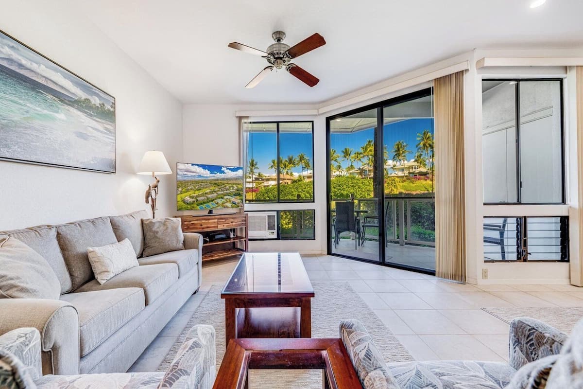 A modern living room with a sofa, coffee table, and TV, a ceiling fan, and a large sliding glass door leading to a balcony with a view of palm trees and houses.