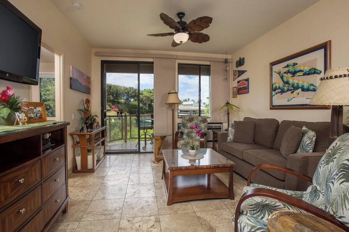 A living room with a television, wooden furniture, a ceiling fan, and a balcony with a view.