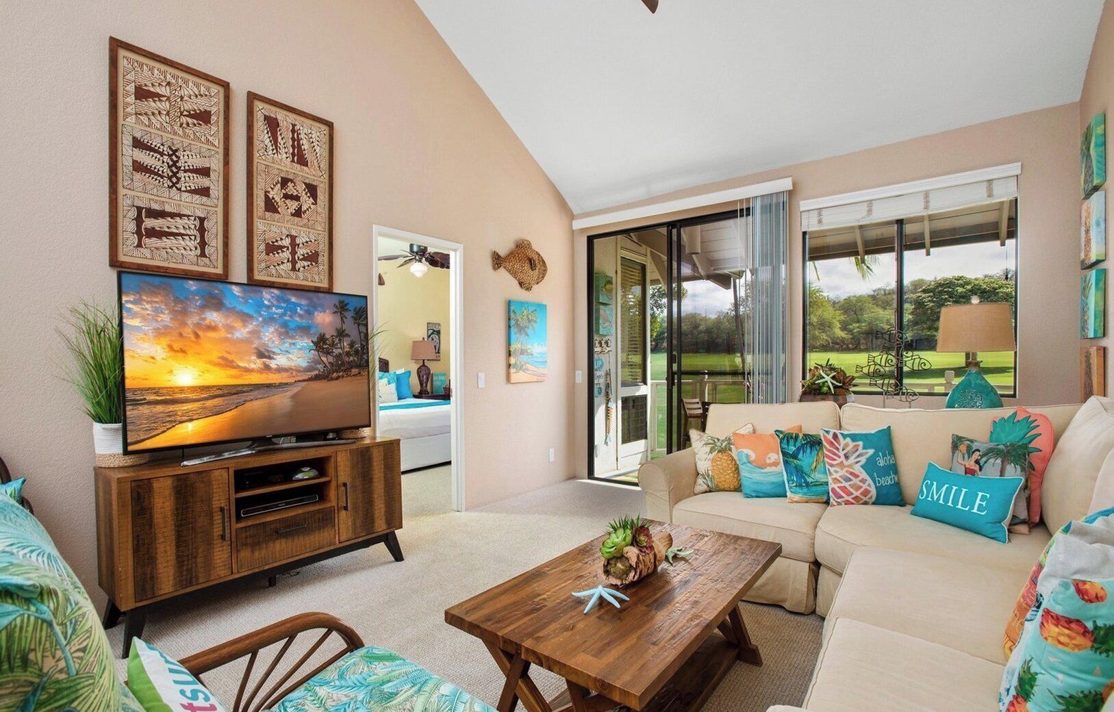 A living room with a TV, wooden entertainment unit, sofa with colorful pillows, and a view of a garden through large sliding doors.