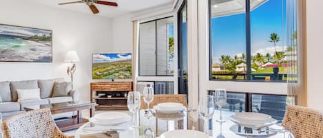 Un espace repas moderne avec une table en verre, des chaises en osier et une vue sur un paysage côtier grâce à de grandes fenêtres.
