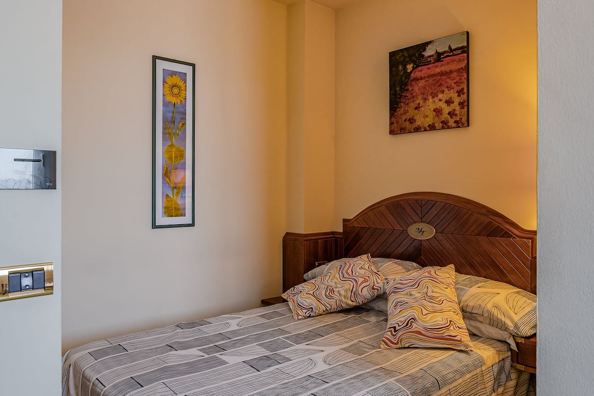 Un dormitorio con cabecera de madera, colcha estampada y dos almohadas. Se observan dos cuadros enmarcados en las paredes.