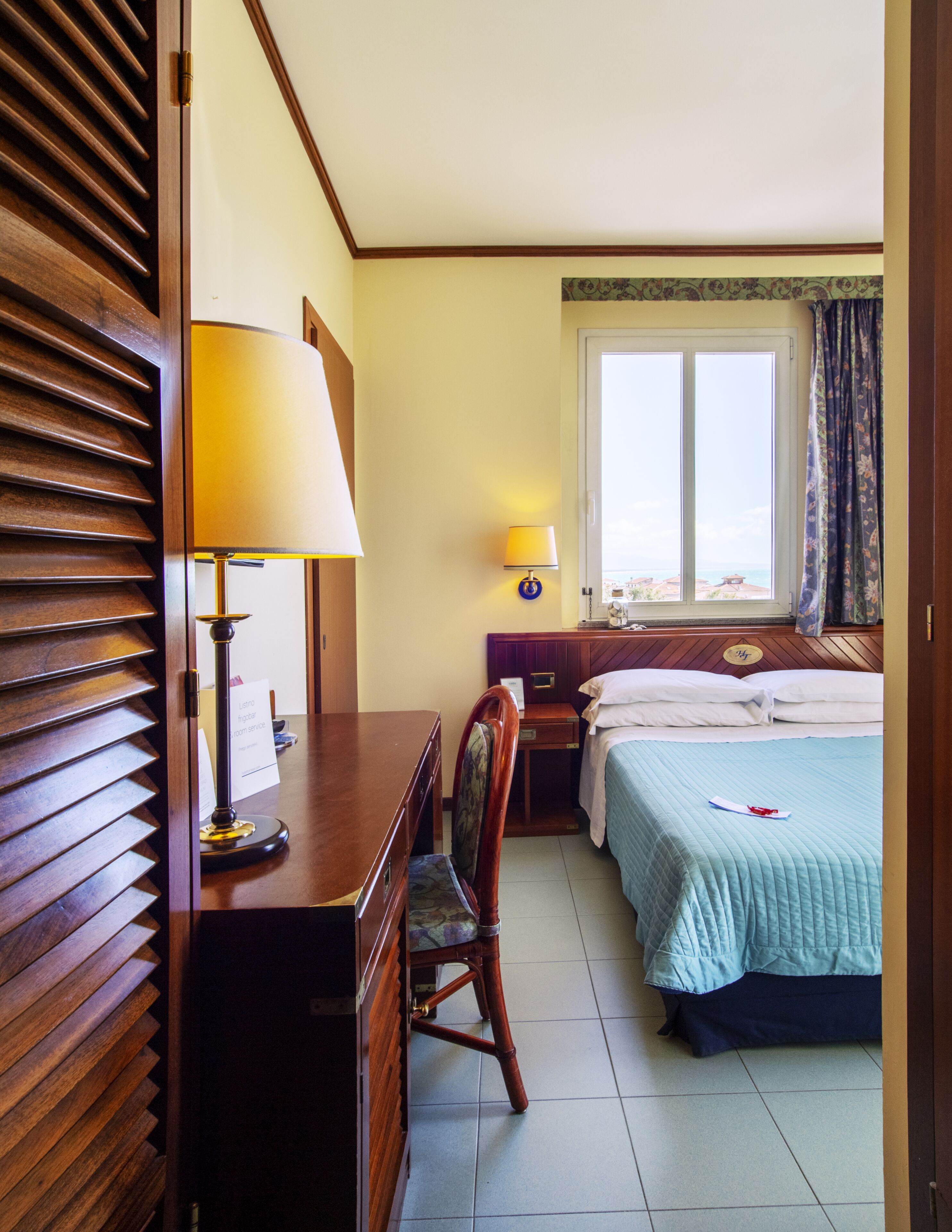 Ein Hotelzimmer mit Holztisch, Stuhl, Bett mit blauer Bettdecke, Nachttisch mit Lampe und einem Fenster mit Vorhängen.