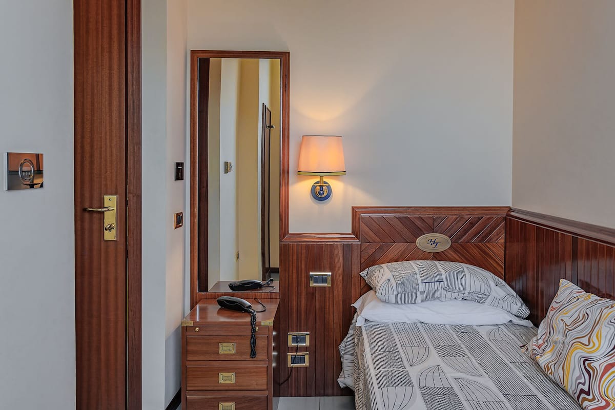 Habitación de hotel con cómoda de madera, teléfono, espejo, cama con ropa de cama estampada y lámpara fijada a la pared.