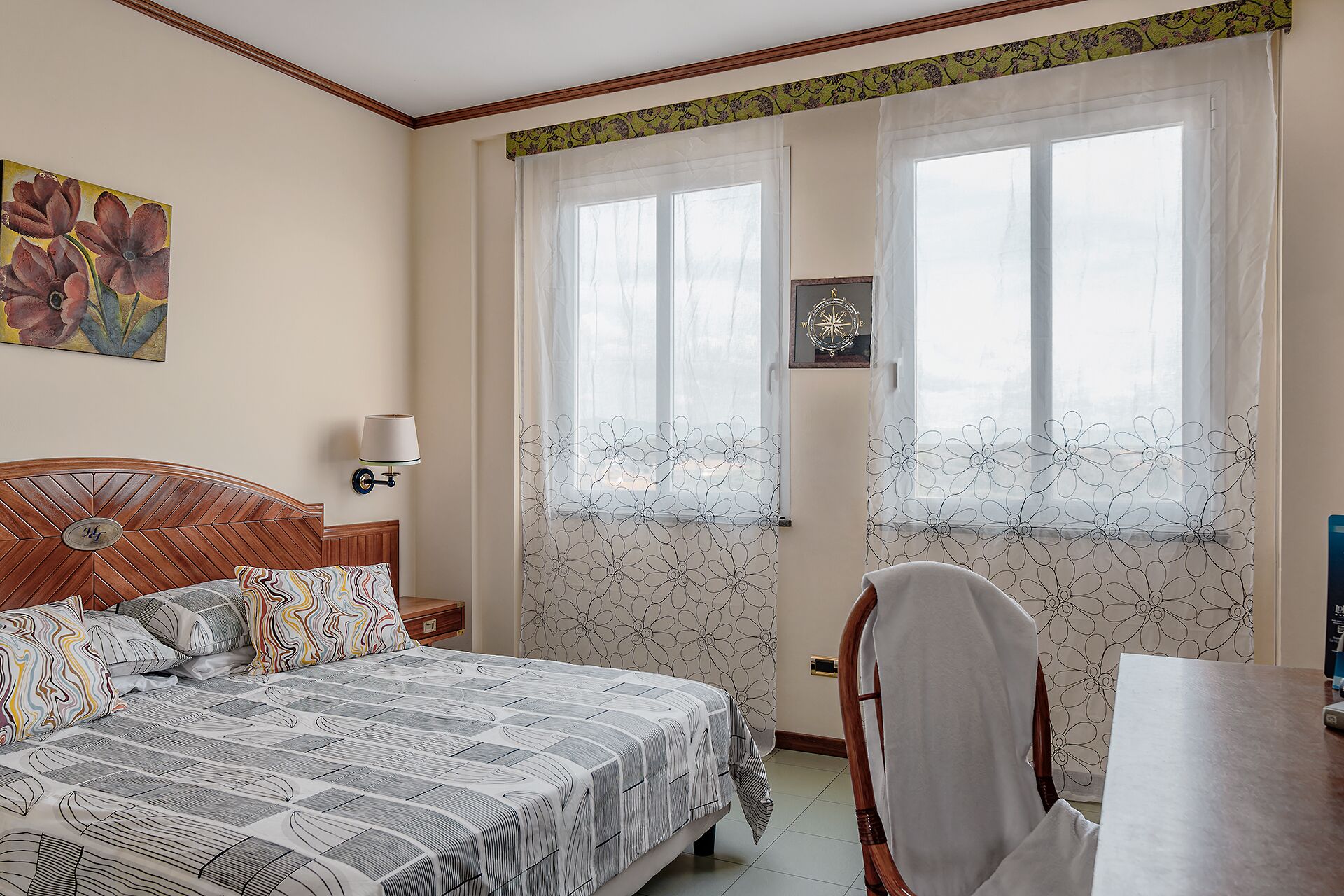 A bedroom with a bed, a nightstand, a chair, and a desk. There are two windows with curtains and a painting on the wall.