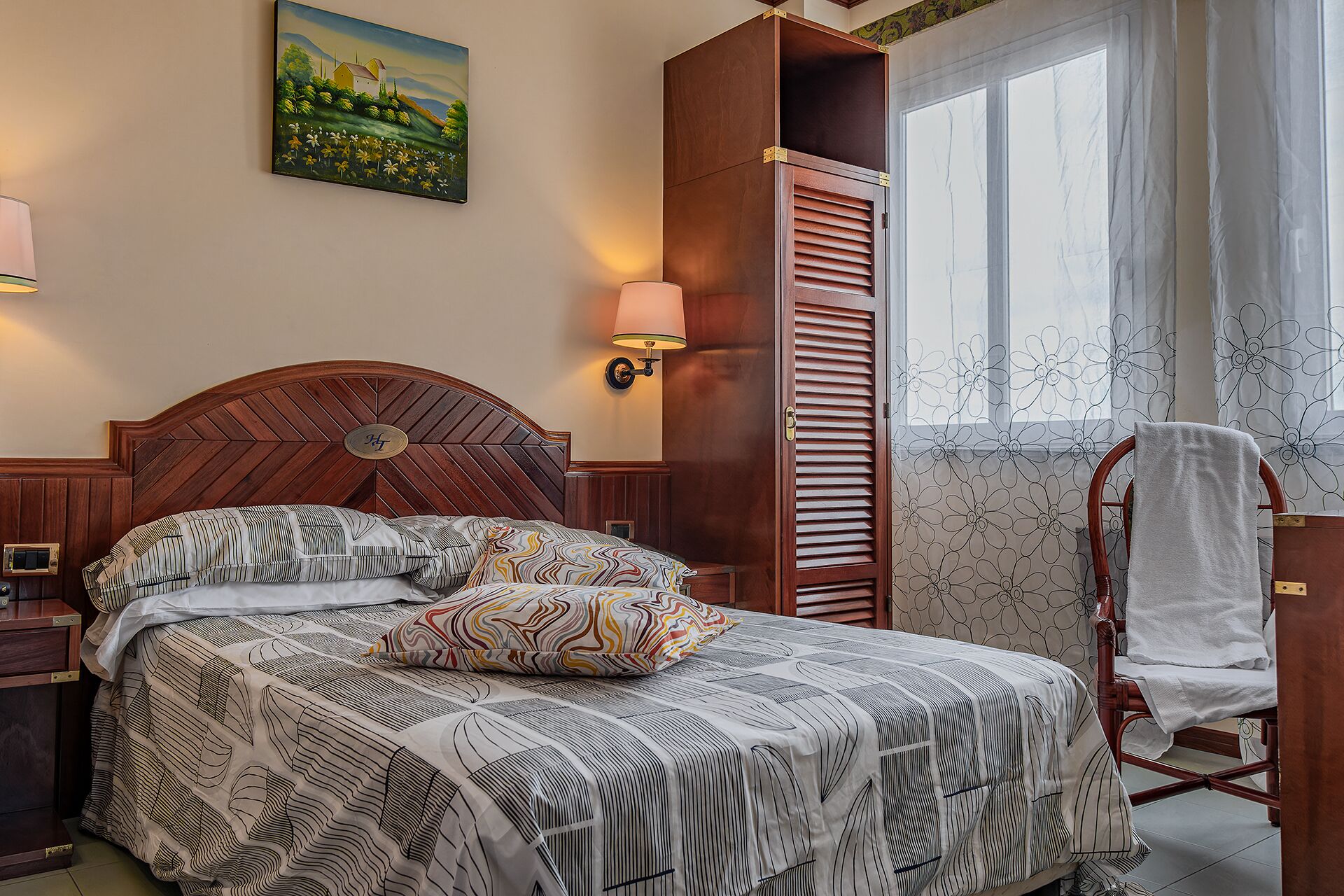 A neatly made bed with patterned bedding, a wooden headboard, a nightstand with a lamp, a wardrobe with a mirror, and a chair with a towel.